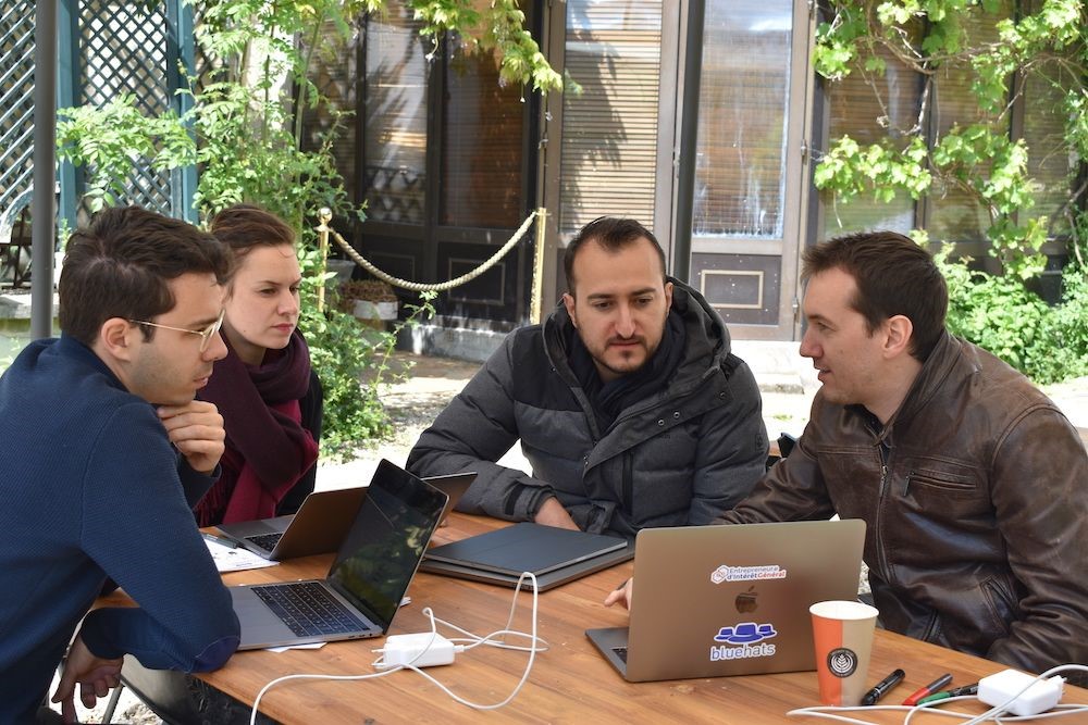 Une femme et trois hommes sont assis autour d'une table, en l'extérieur, sur laquelle on voit plusieurs ordinateurs. Ils discutent en regardant quelque chose sur un des ordinateurs.